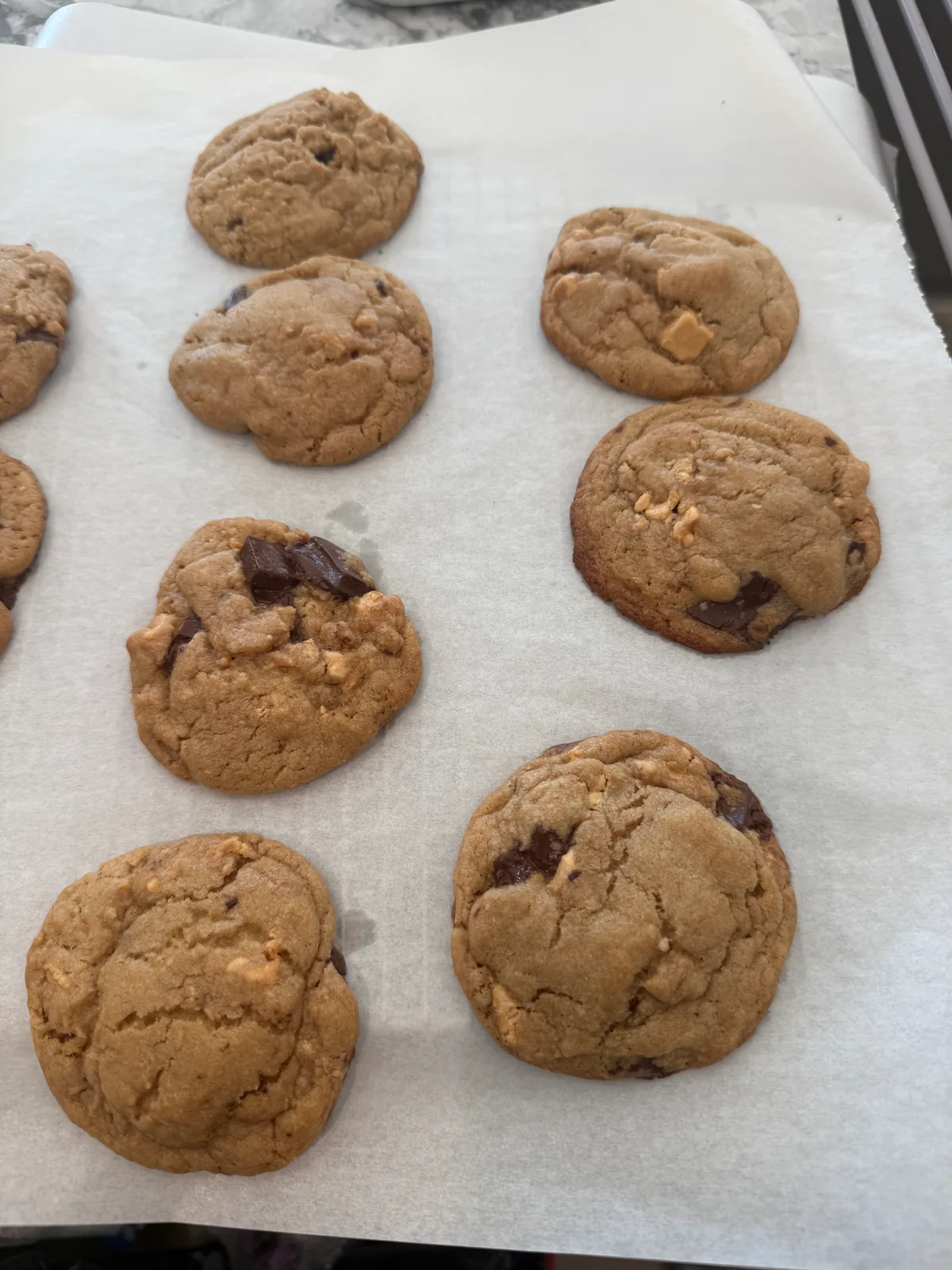 Golden-brown cookies with cracked tops and dark and pale chunks rest on white parchment paper on a baking tray.