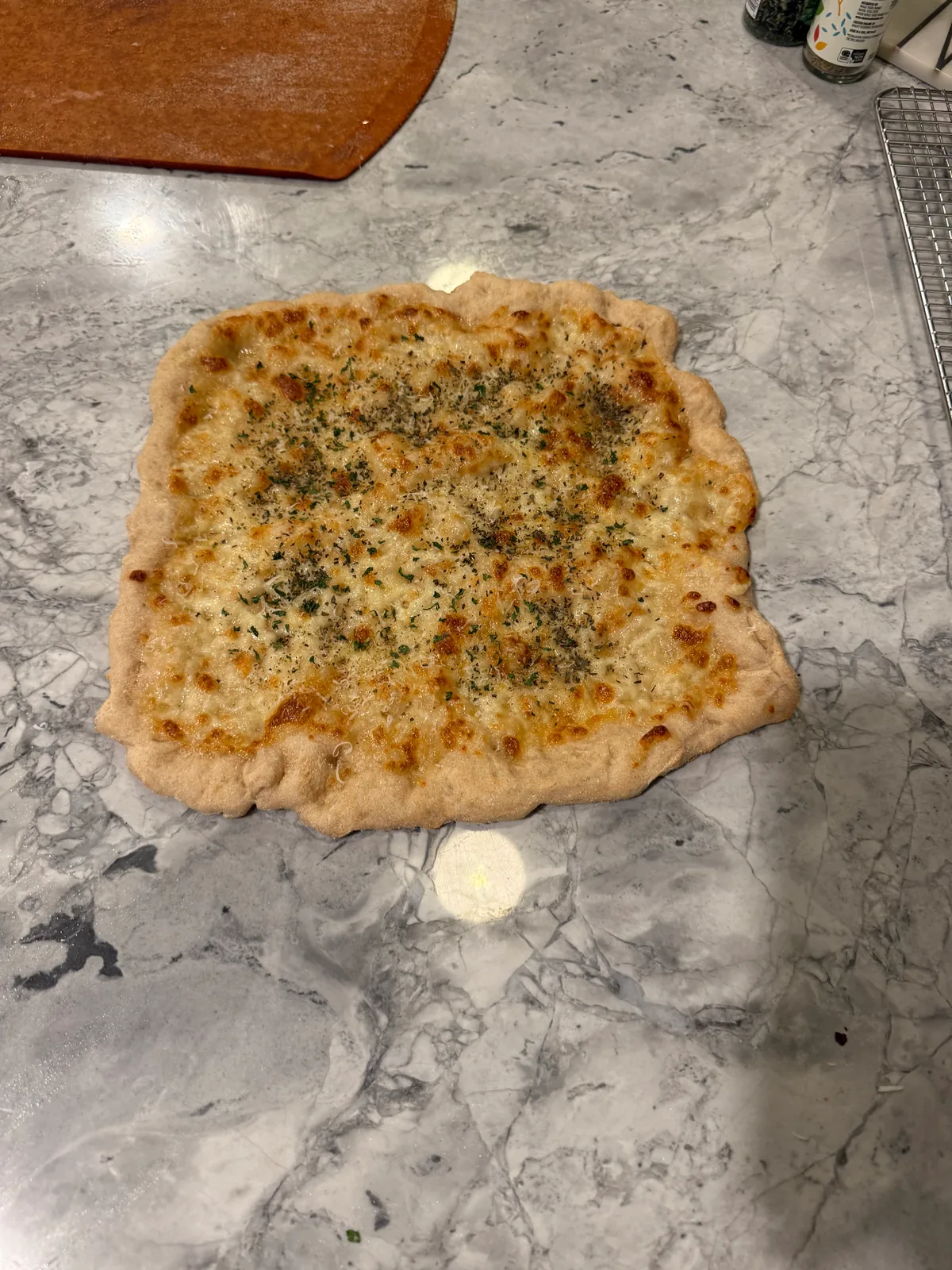 An irregular square cheese-and-herb flatbread pizza with browned, bubbly cheese and a thick pale crust on a gray marble countertop.