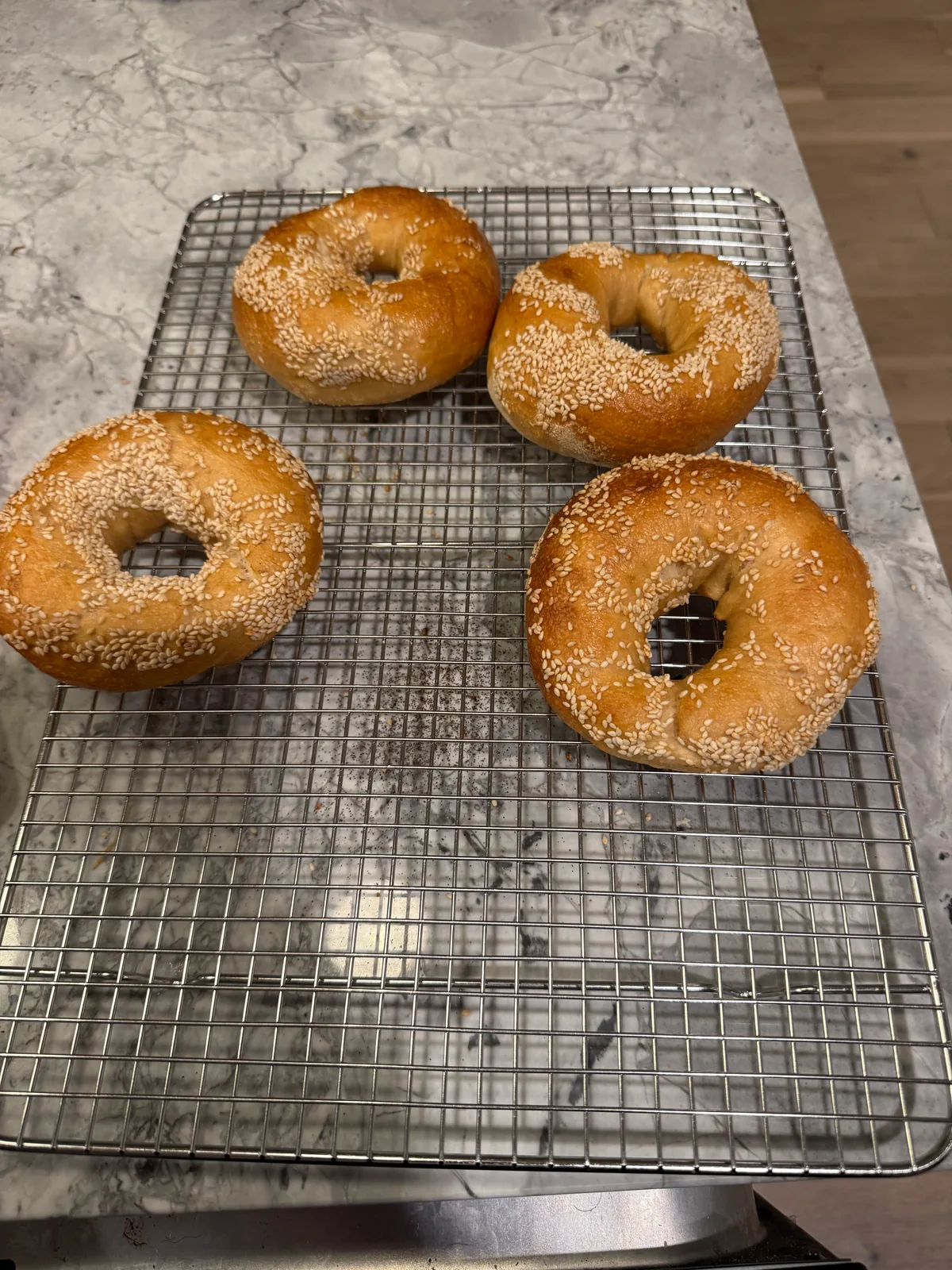 Four golden-brown, sesame-covered bagels rest on a rectangular wire cooling rack on a gray marble-patterned countertop.