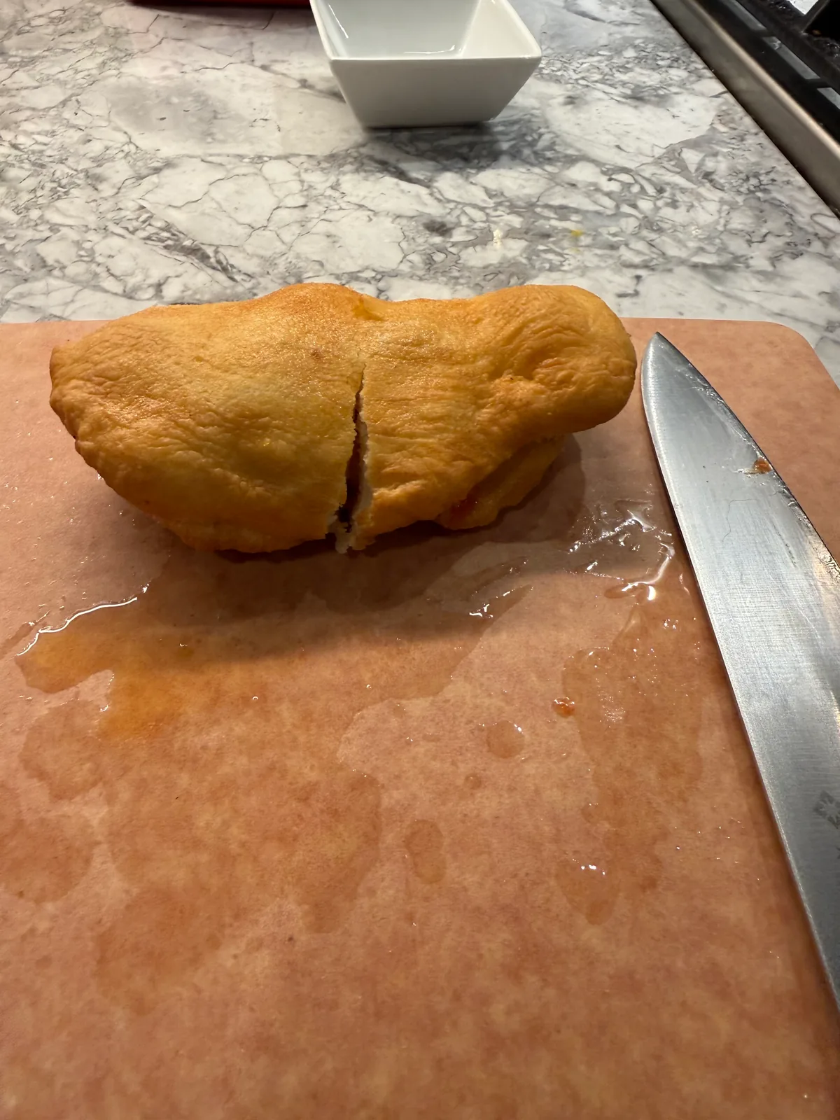 A golden-brown dough pocket cut across the middle rests on a wet pink cutting board beside a large knife, with a white bowl on the marble-patterned counter behind it.