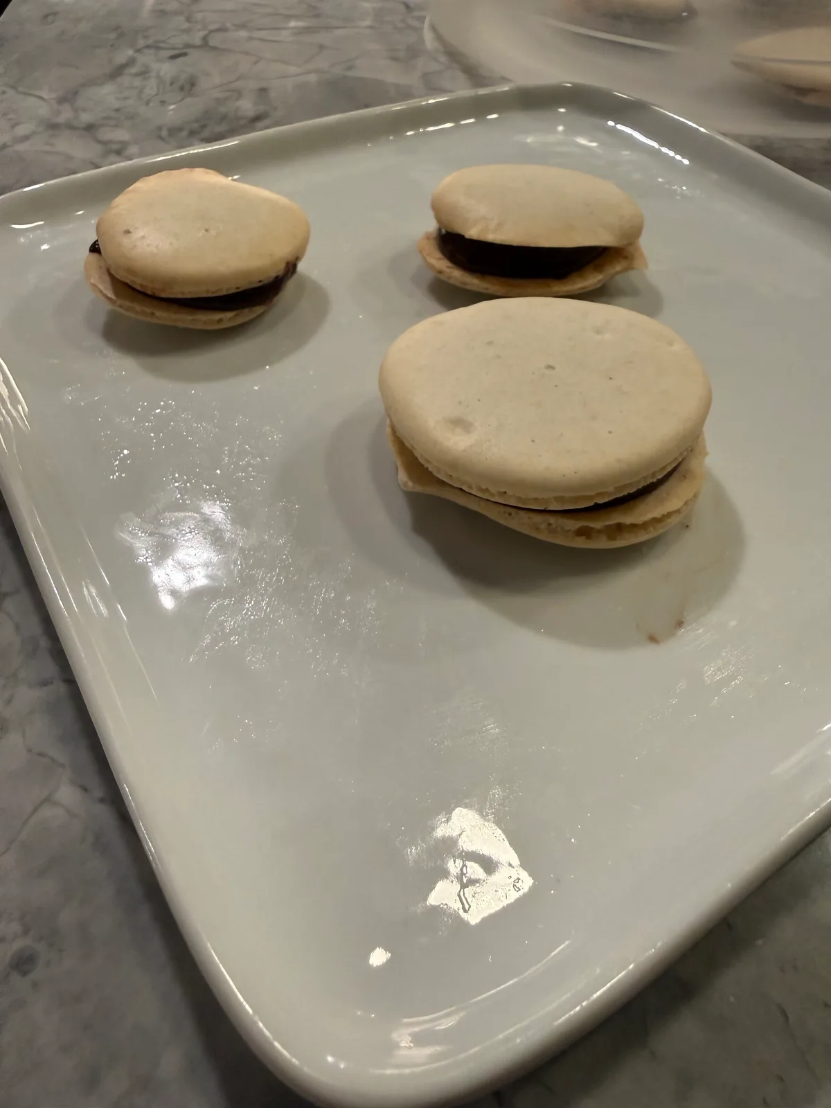 Three pale macarons with dark filling on a glossy rectangular white plate on a gray marble countertop.