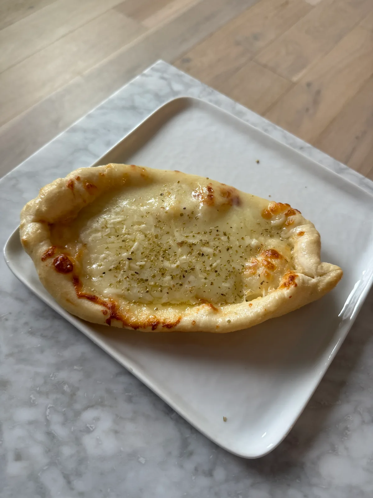 Boat-shaped flatbread filled with melted cheese, browned in spots and sprinkled with herbs, on a white rectangular plate atop a marble table.