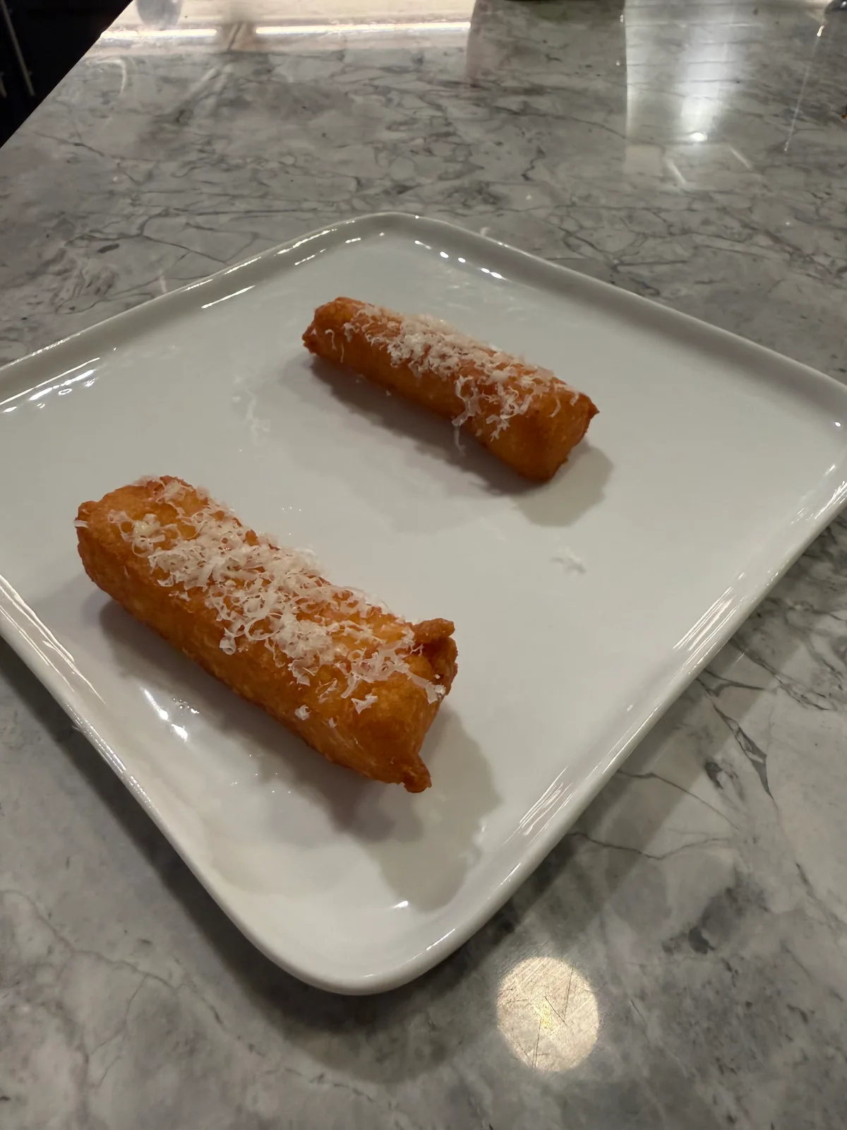 Two golden-brown fried batons topped with fine white cheese shavings on a square white plate on a gray marble counter.
