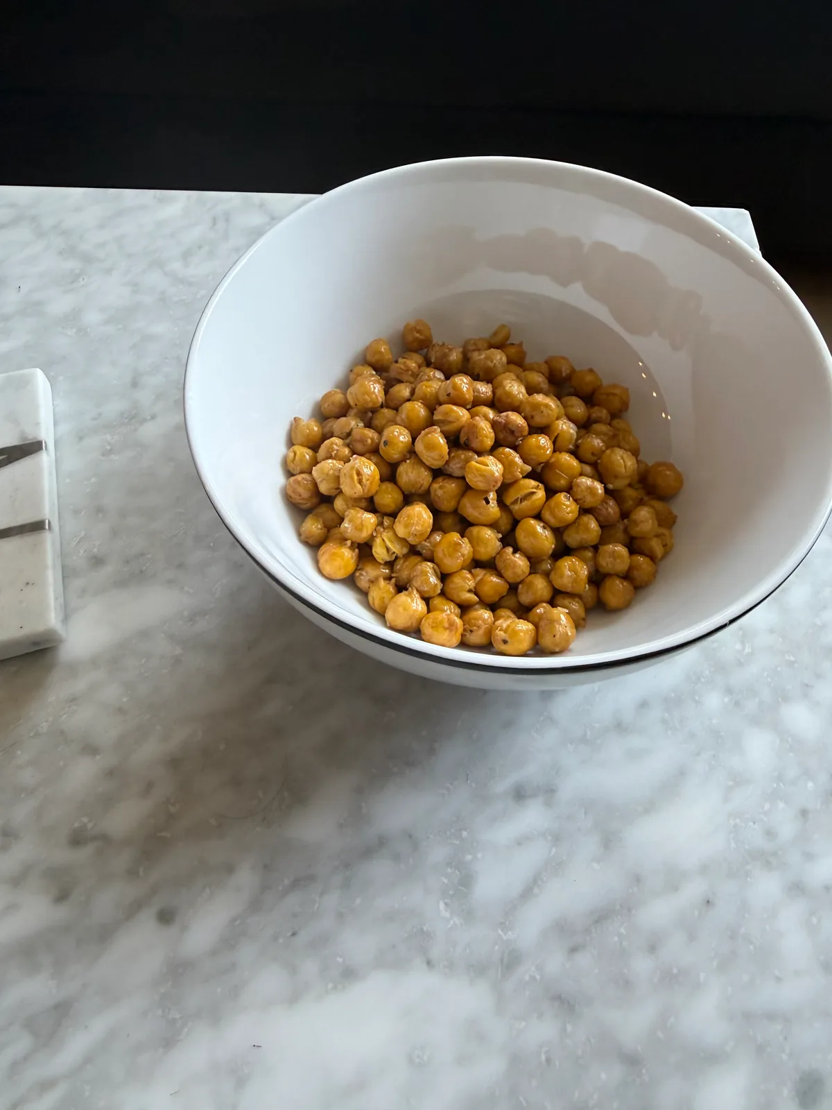 A white bowl filled with golden-brown roasted chickpeas sits on a pale marble countertop.