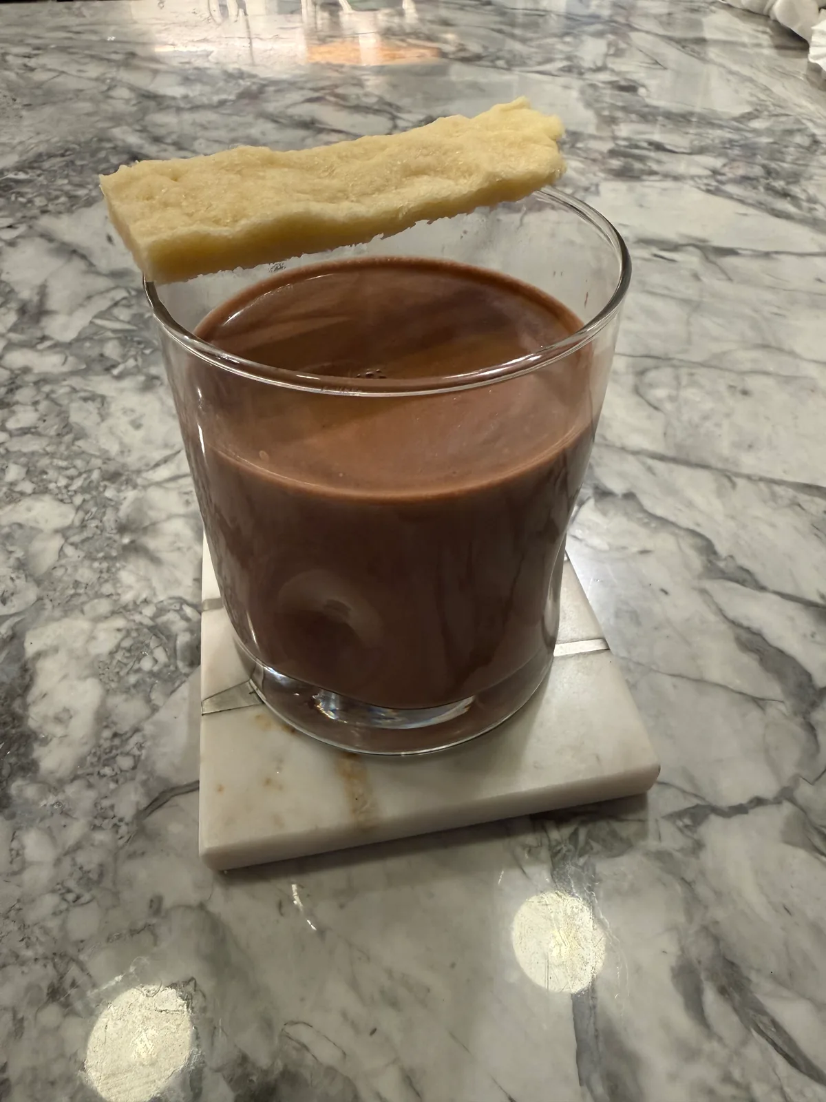A glass of dark brown liquid with a pale rectangular biscuit resting across its rim, on a square stone coaster on a gray marble countertop.