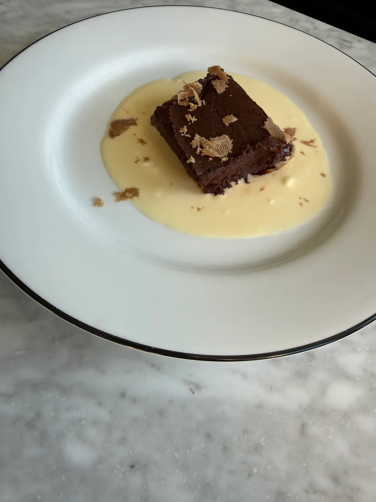 A square of dark chocolate cake topped with beige flakes sits in a pool of pale yellow sauce on a white plate with a dark rim.